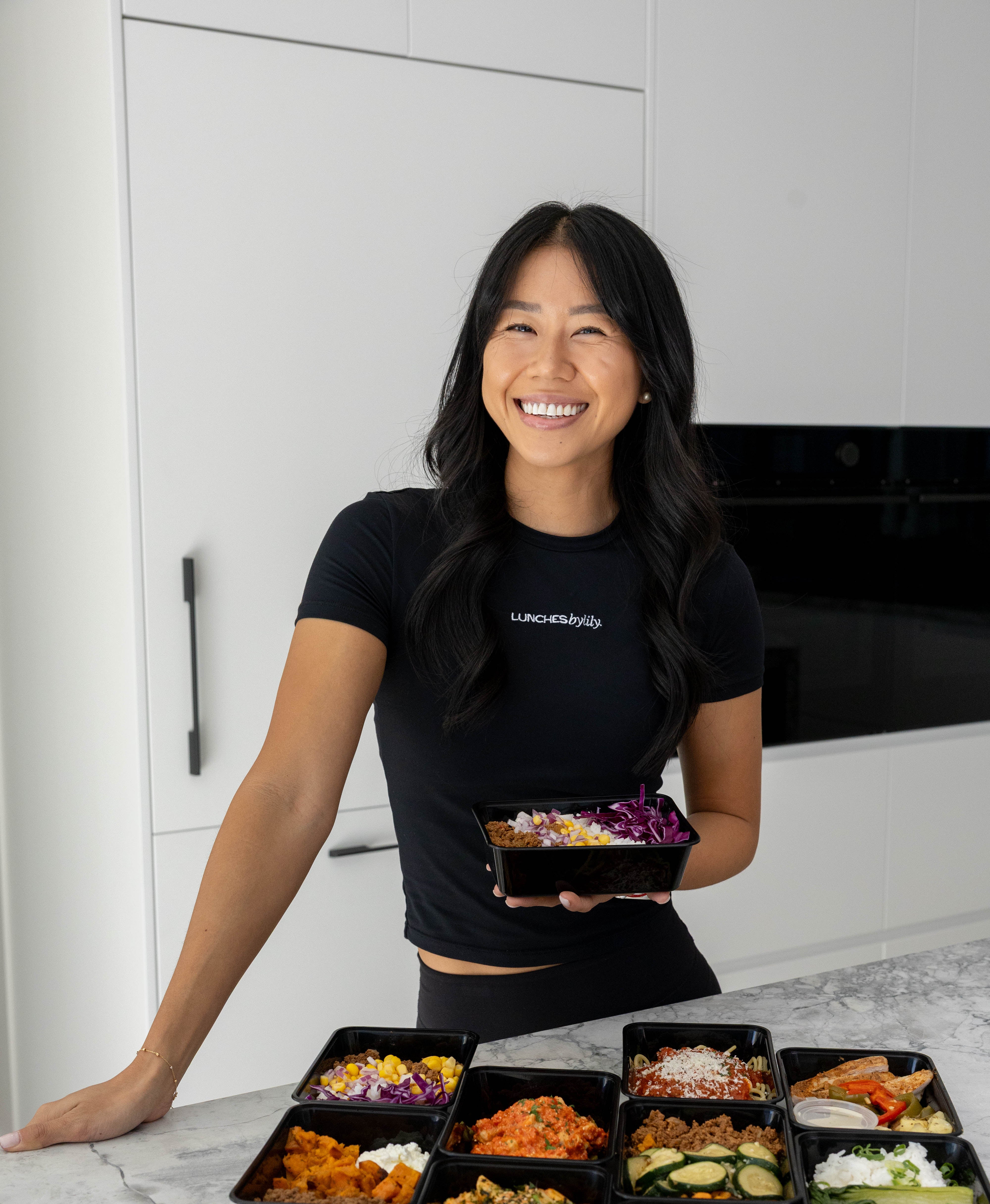 Woman holding a container of food with multiple prepared meals on a kitchen counter.